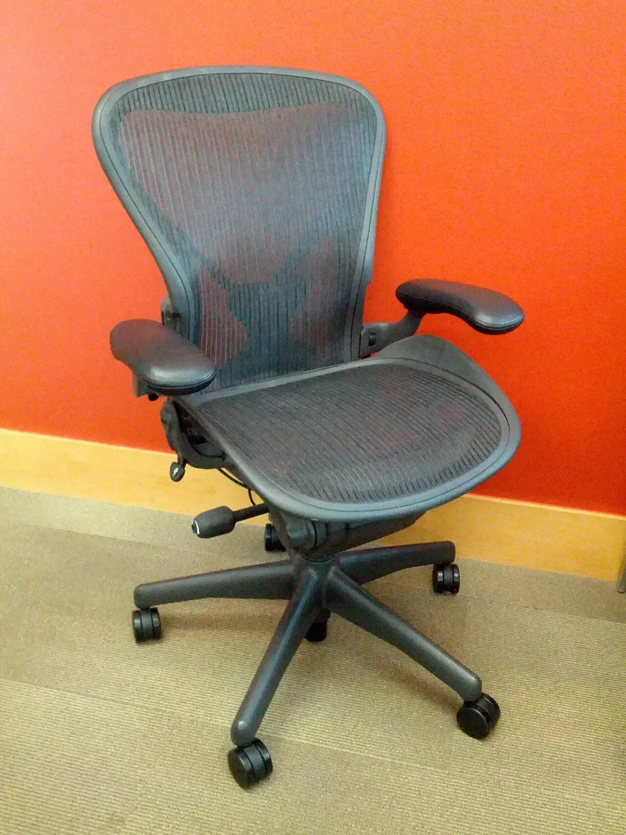 A Herman Miller Aeron Classic office chair with dark mesh and padded arms, standing in front of an orange wall