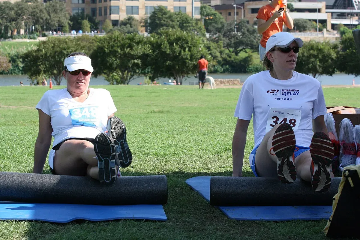 Two runners sitting on mats on the grass, rolling their hamstrings on long foam rollers after a race