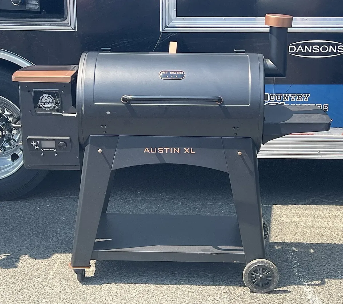 A black Pit Boss Austin XL pellet grill with copper accents, a side hopper with a digital controller, and a bottom shelf, parked outdoors